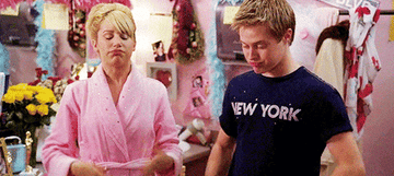 Mujer y hombre jóvenes en escena de película, ella con bata rosa y él con camiseta con texto &quot;New York&quot;
