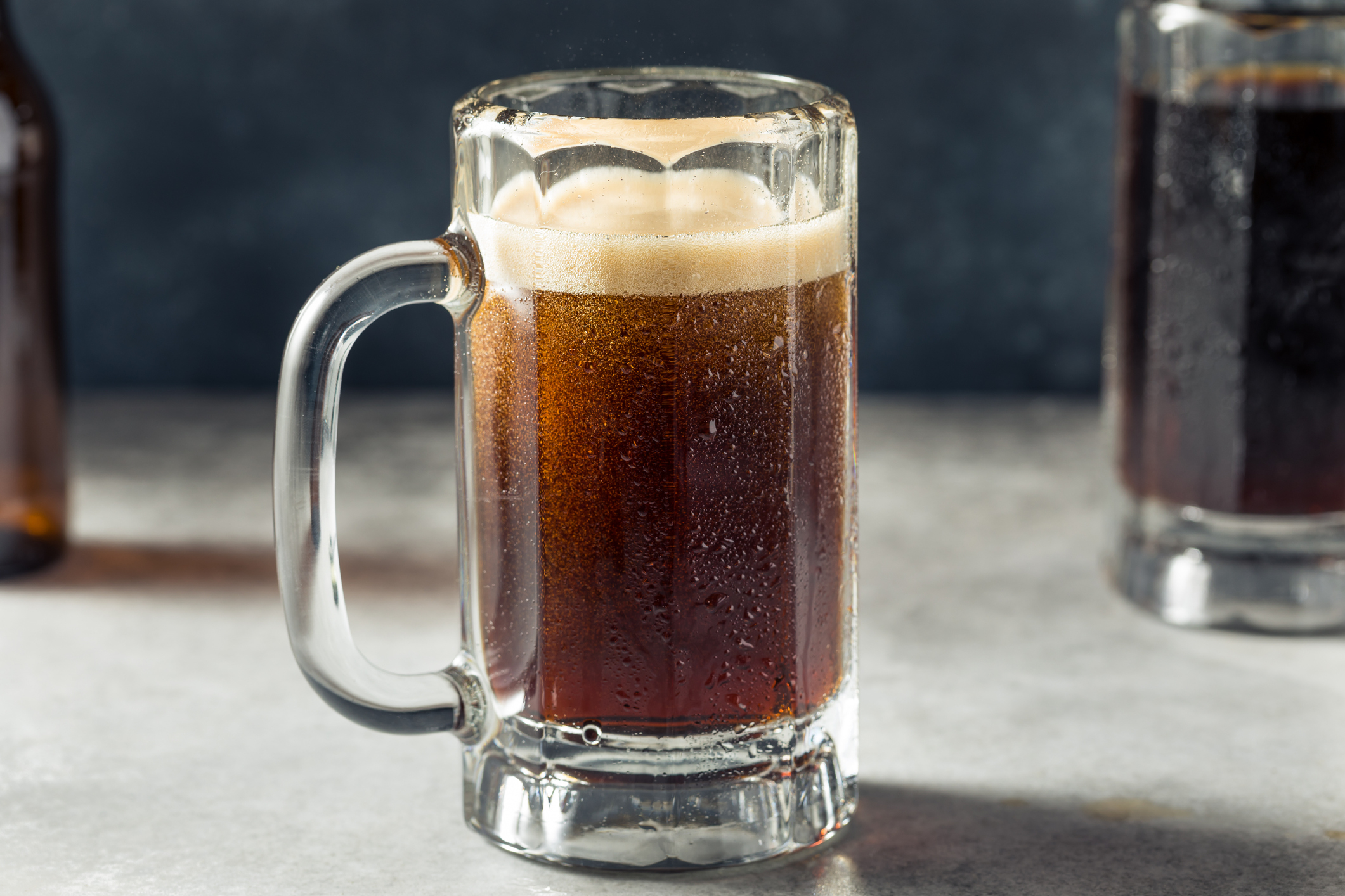 Mug of beer with frothy head on table, background with dark bottle and glass
