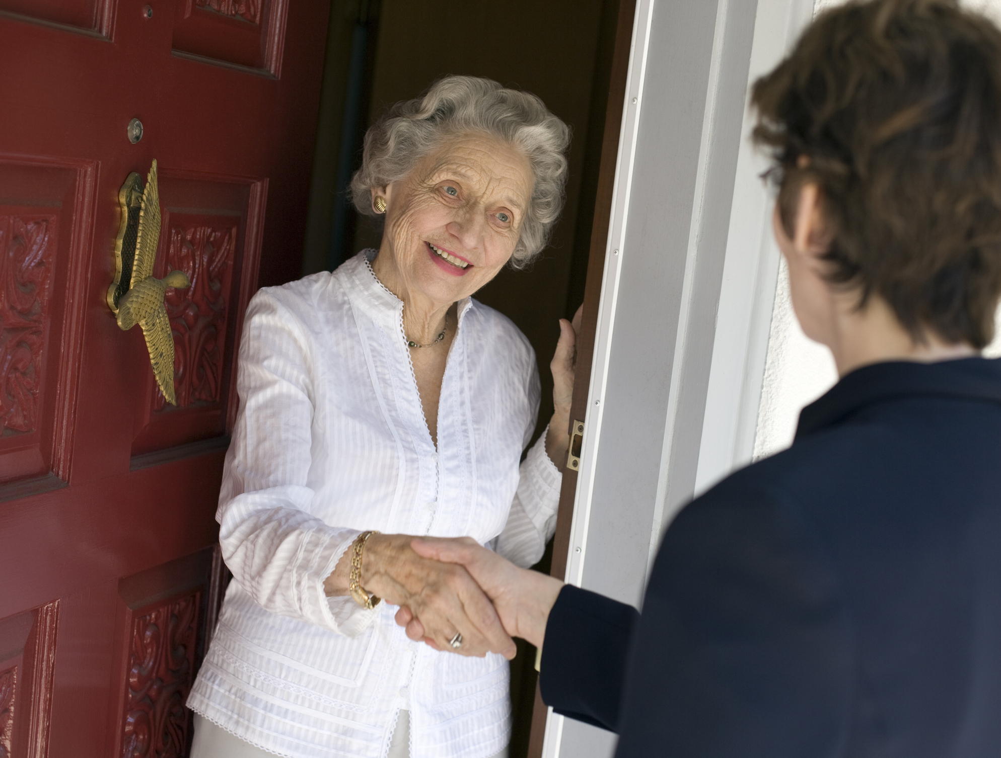 Older woman greets visitor at her doorway with a handshake and a smile