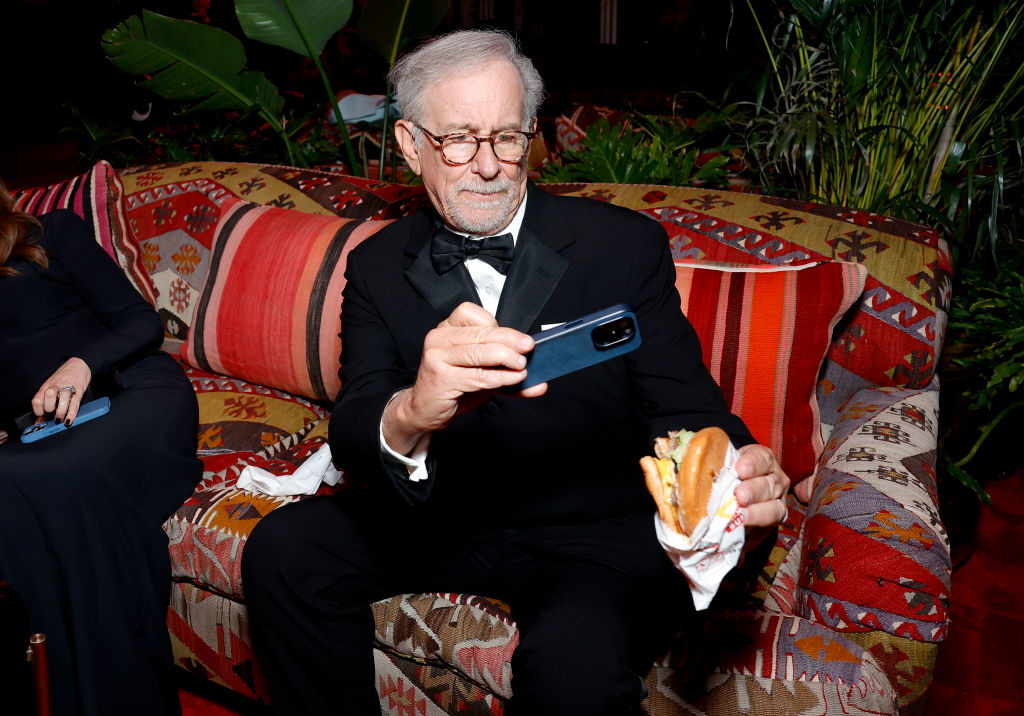 Steven Spielberg sitting on a couch, eating a sandwich, and looking at his phone