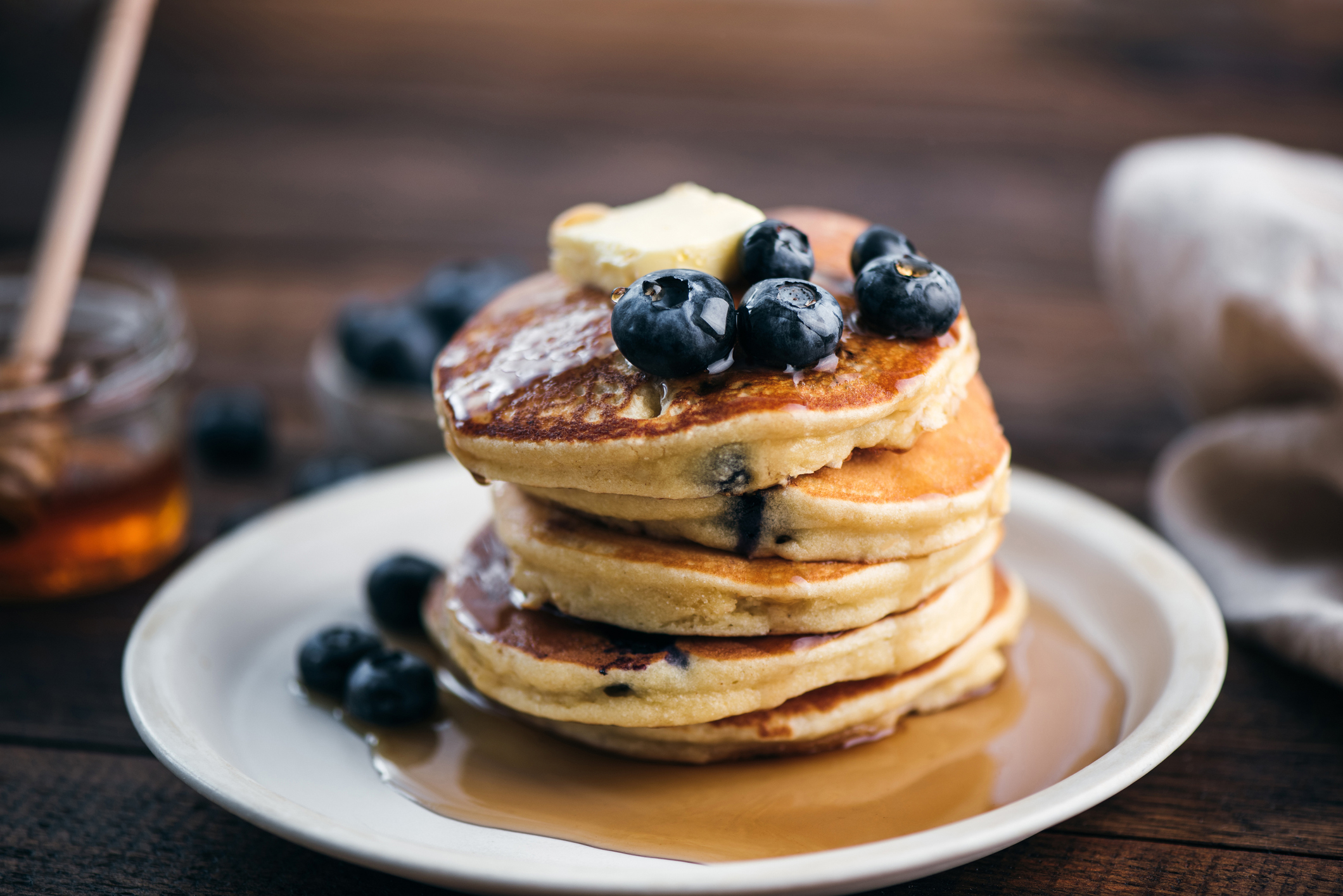 Stack of pancakes with blueberries and a pat of butter, honey on side, on a wooden surface
