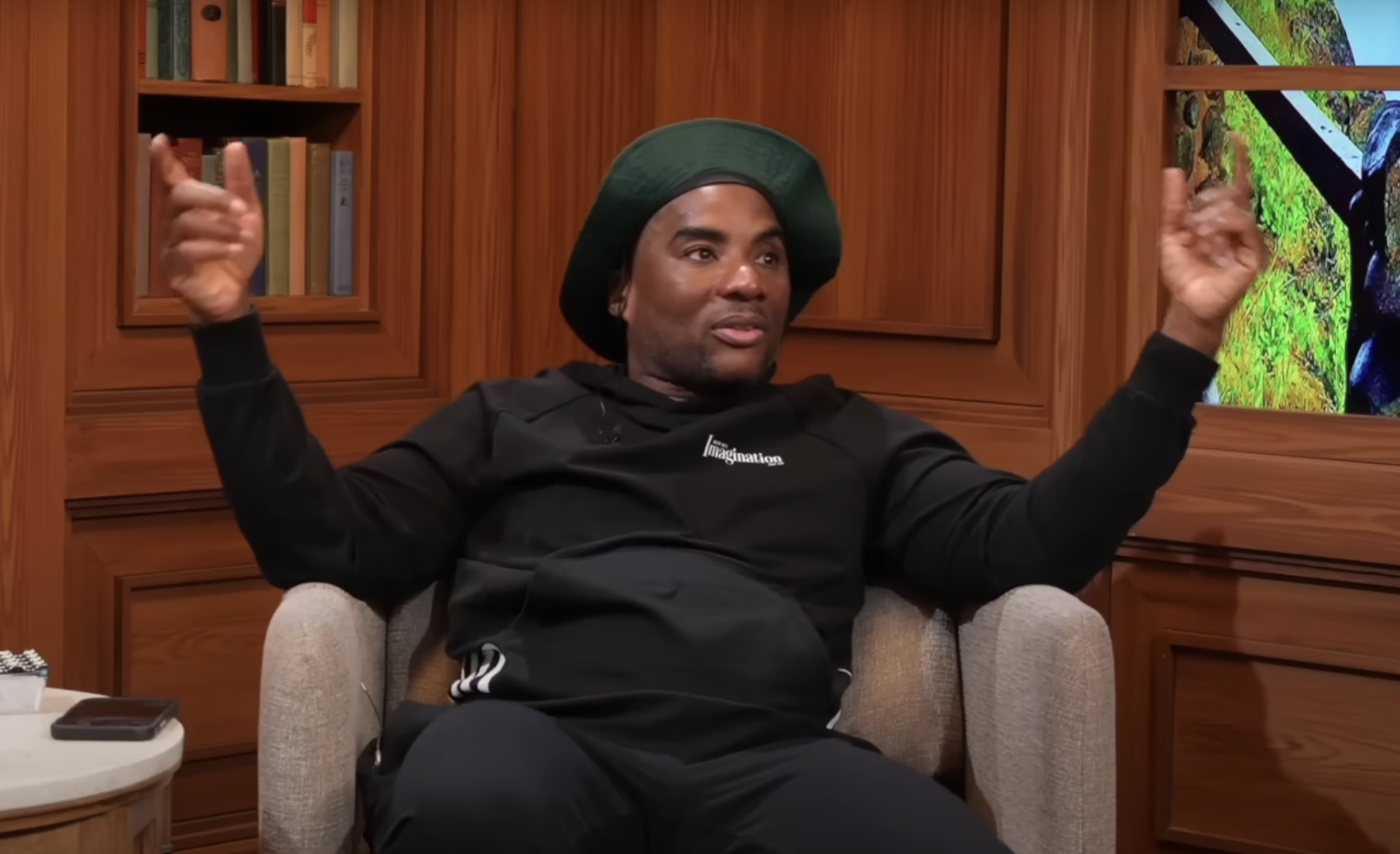 Charlamagne gesturing with hands up, wearing a beret and hooded sweatshirt, sitting indoors