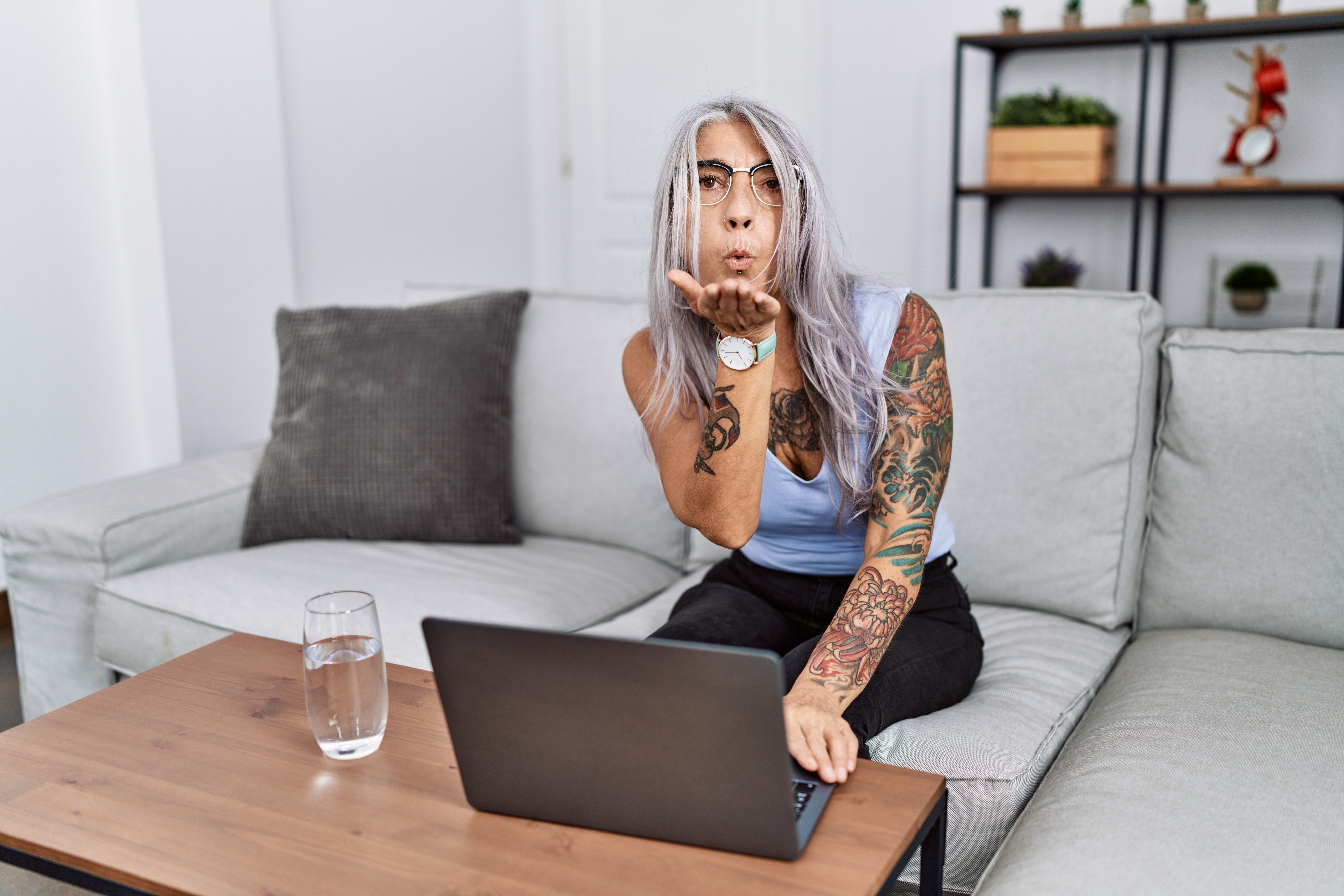 Person with tattoos sitting at a laptop, sending a blowing kiss gesture toward the camera