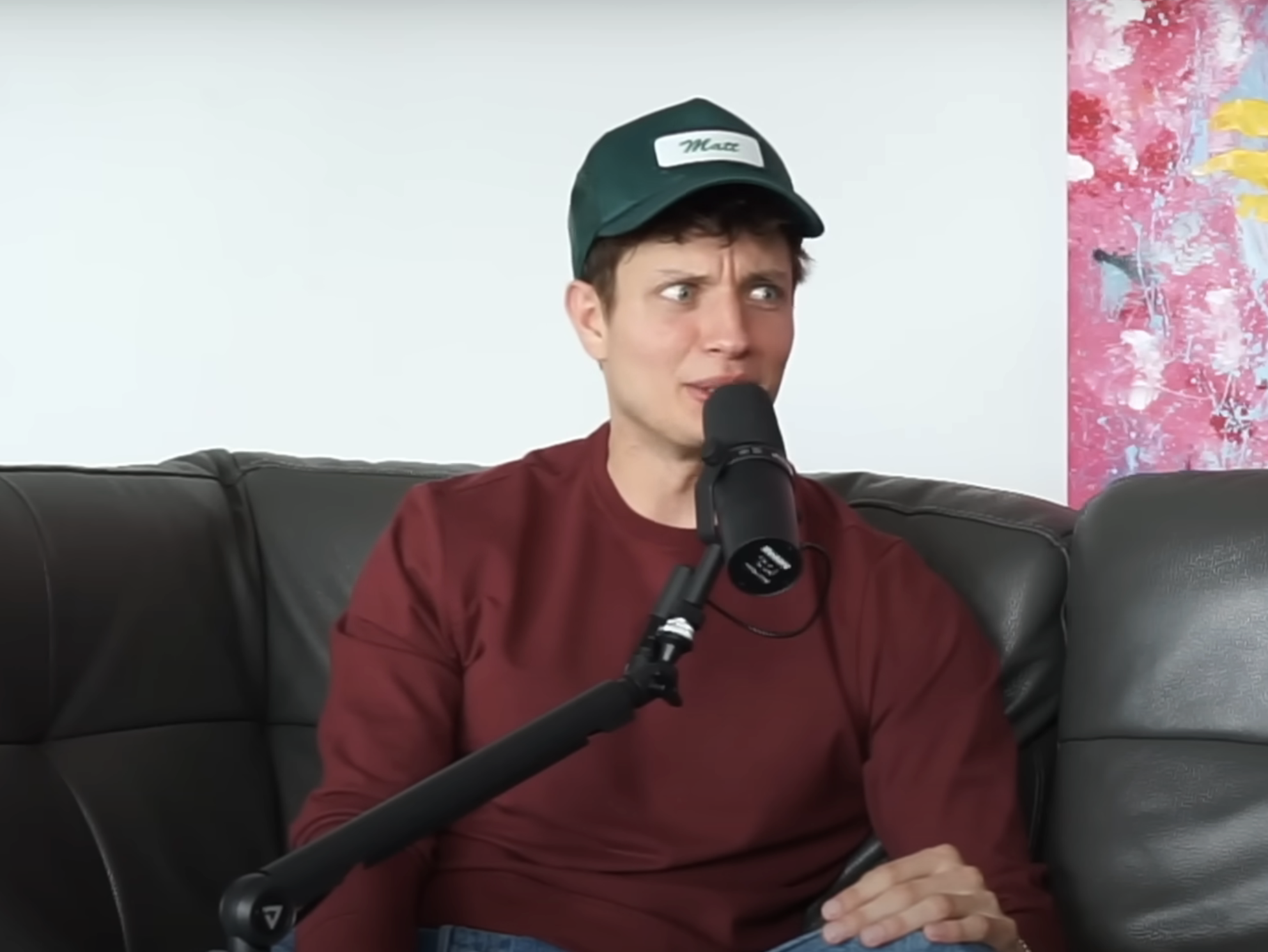 Matt in cap and shirt speaking into a microphone while seated on a couch