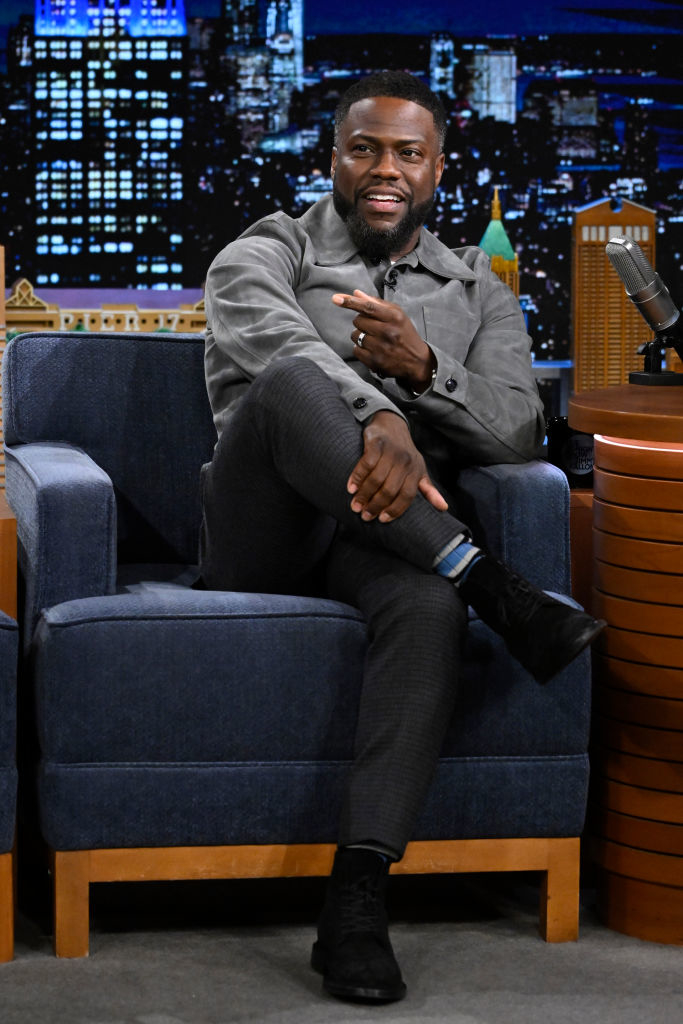 Kevin Hart wearing a stylish jacket, seated with crossed legs, gesturing during a talk show interview