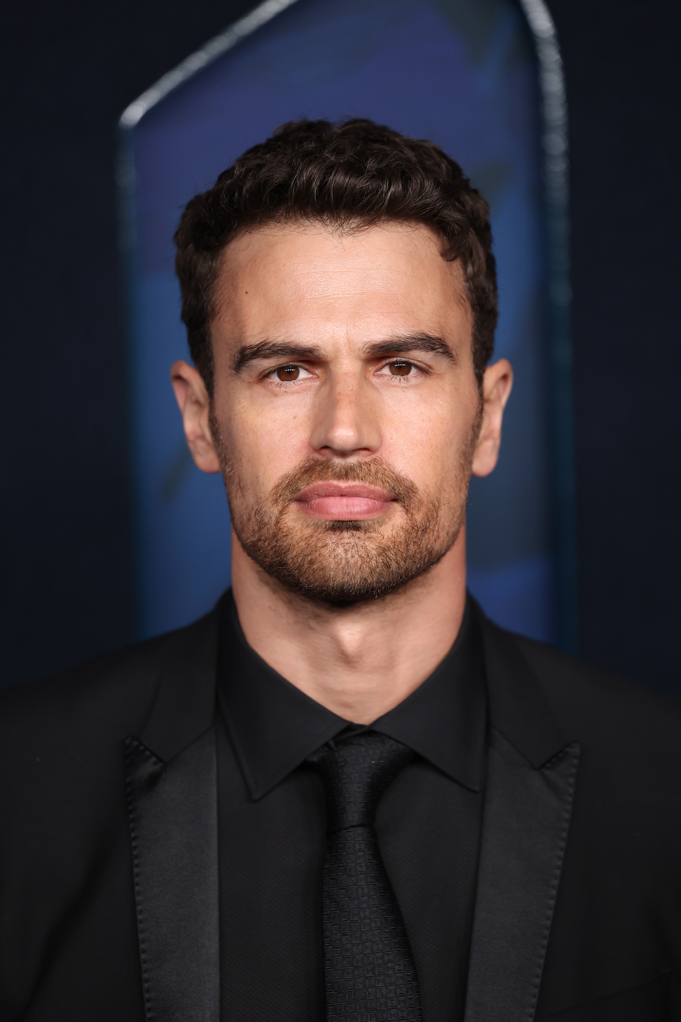 Theo in black suit and tie at an event, serious expression