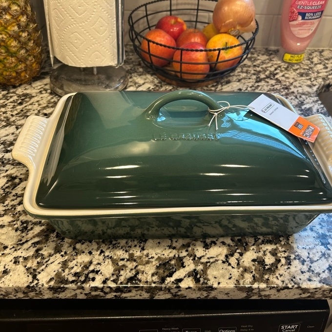 Green ceramic baking dish with lid on a kitchen counter, price tag attached, surrounded by fruits and a paper towel roll