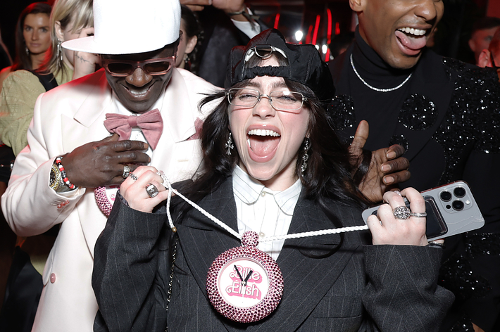 Three exuberant individuals at an event, one in a white suit, another in a black embellished outfit, and a person in the center with a quirky hat and glasses