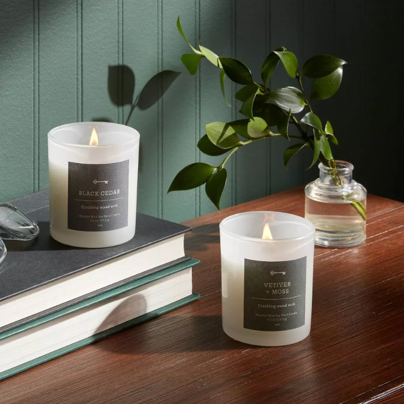 Two lit scented candles on a table, one labeled Black Cedar and the other Vetiver Moss, beside stacked books and a plant