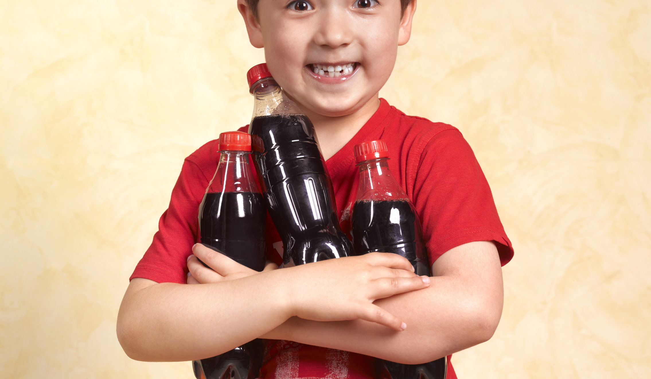 Child smiling while holding three soda bottles, standing against a yellow backdrop