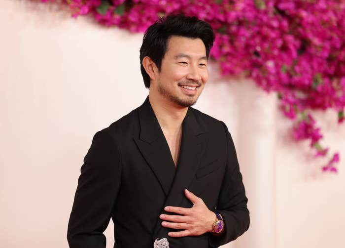 Simu Liu in a suit sans shirt smiling at an event
