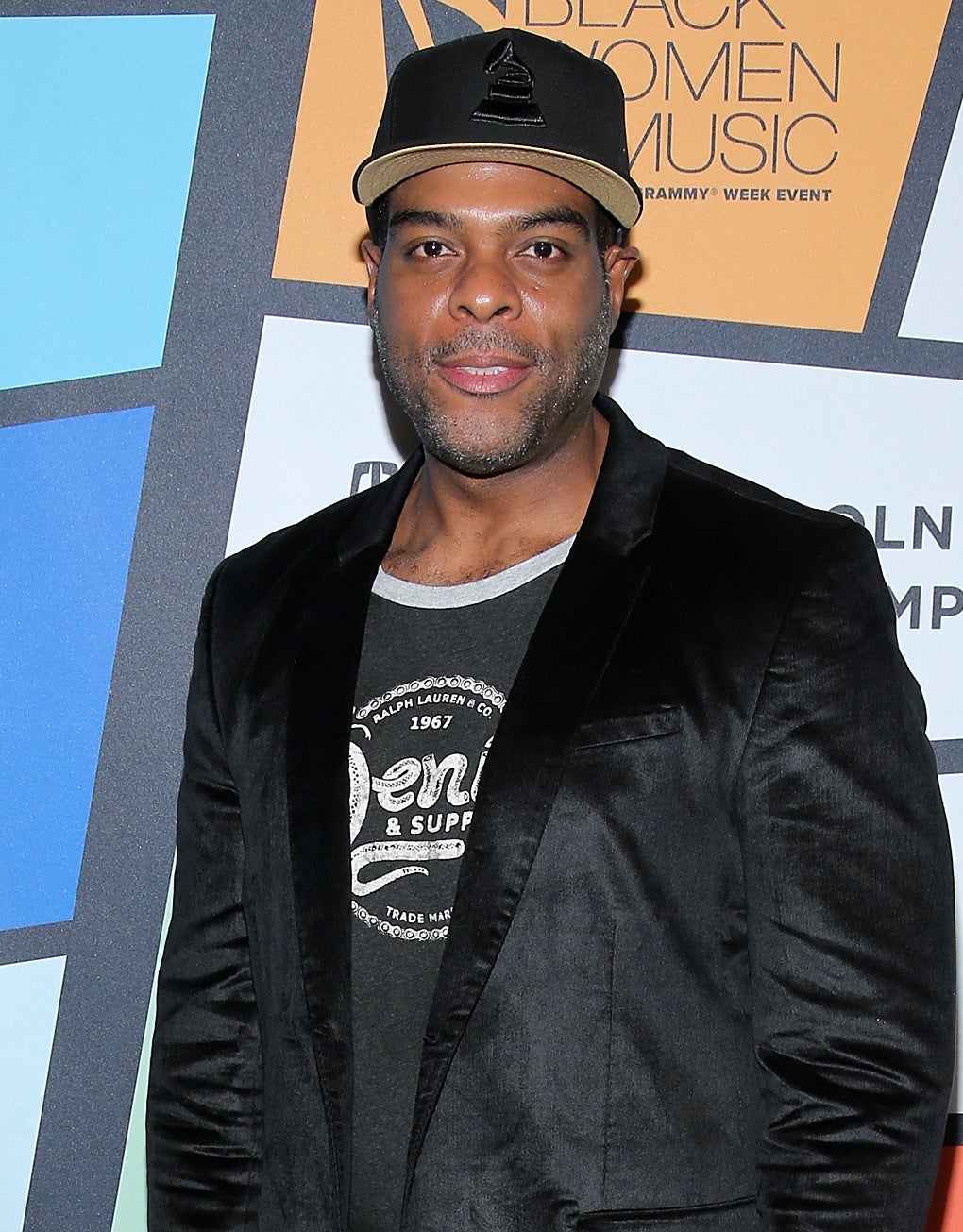 Troy Winbush posing for a photo at a media event wearing a cap, jacket, and graphic tee