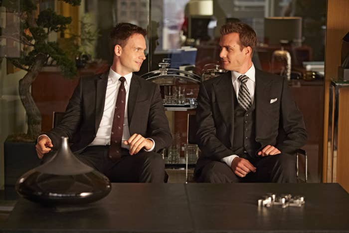 Two characters from a TV show sitting in a office, looking at each other with subtle smiles, dressed in suits