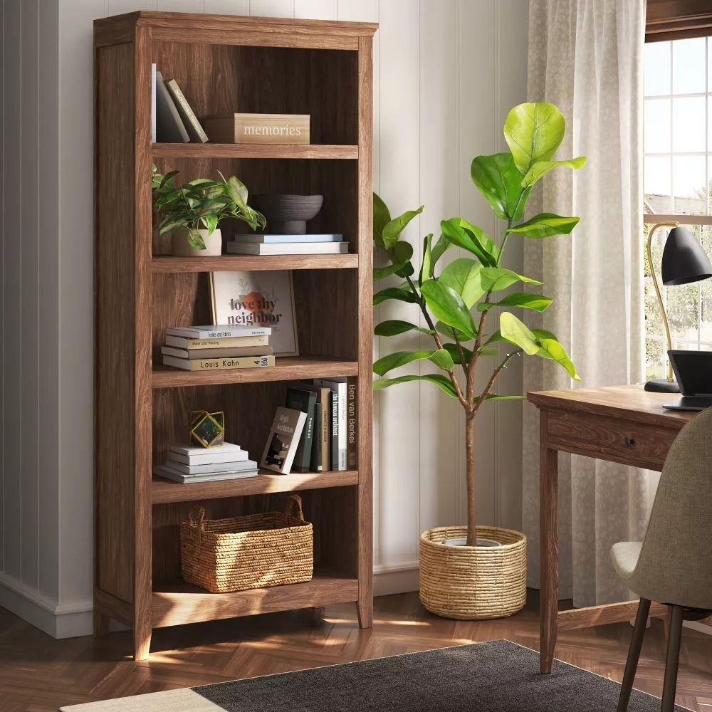 A wooden bookshelf with plants, books, and a basket beside a desk in a home office setup