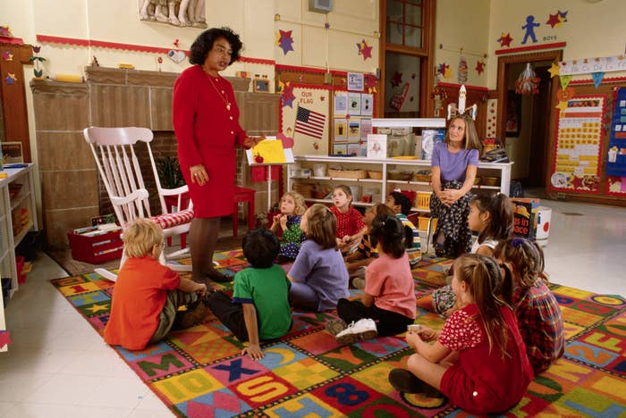 A teacher standing in a classroom, with children sitting on the floor looking at her. She&#x27;s holding a book