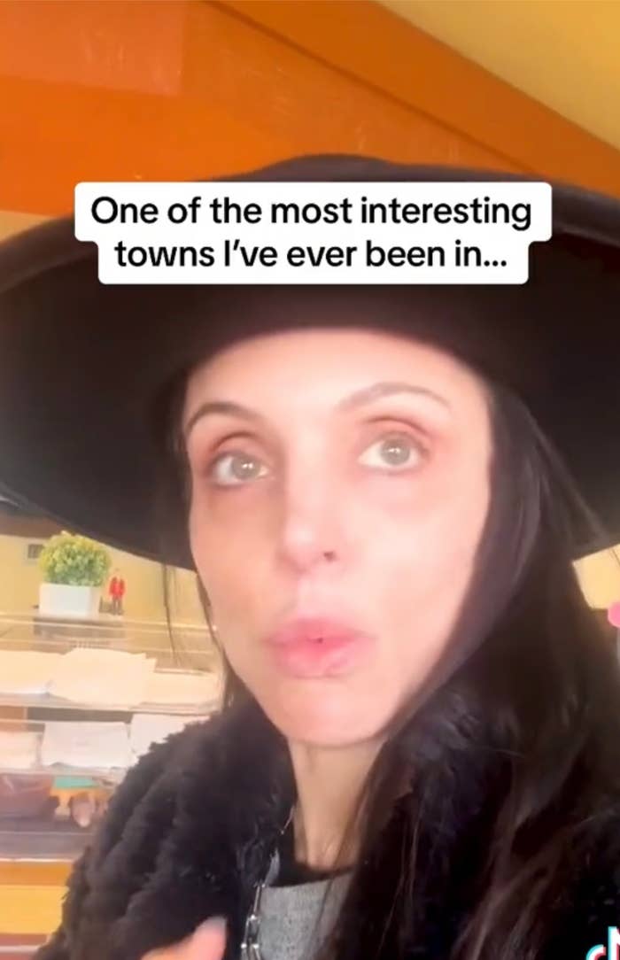 Woman in hat with surprised expression, text overlay: "One of the most interesting towns I’ve ever been in…"
