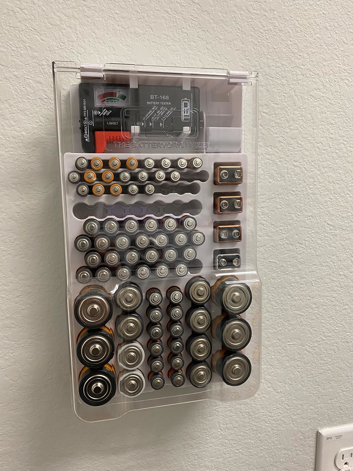 A wall-mounted clear battery organizer stocked with various sizes of batteries