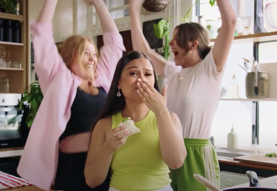 Three women in a kitchen celebrating, one laughs covering her mouth, others cheer with raised arms