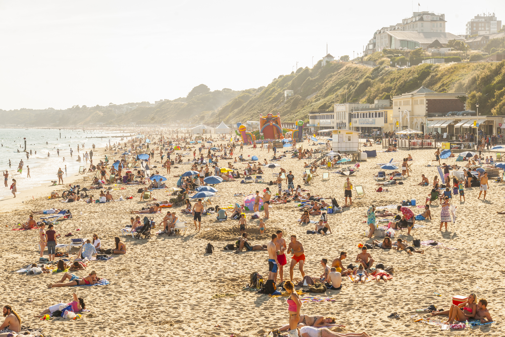 Busy beach scene with people sunbathing and walking, including umbrellas and a backdrop of buildings on a hill