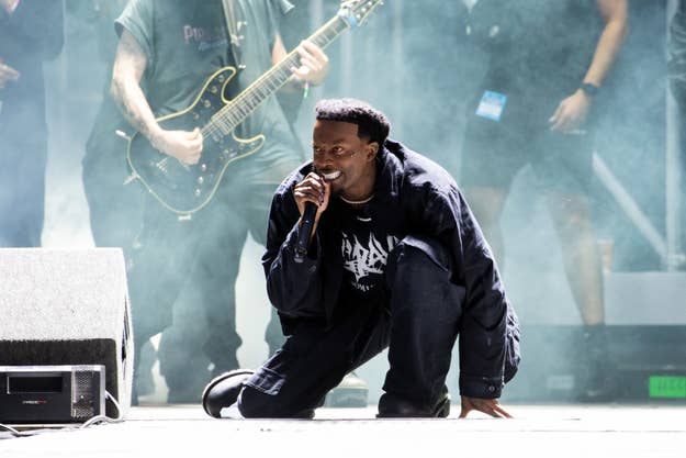 Playboi Carti kneeling on stage with microphone, performing live with guitarist in background