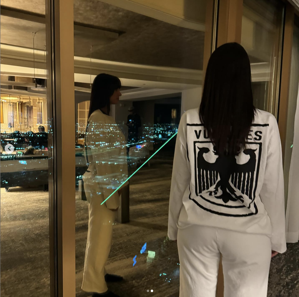 Woman in a white jacket with &quot;VOTES&quot; text at the back looks at her reflection in a window overlooking a city night view
