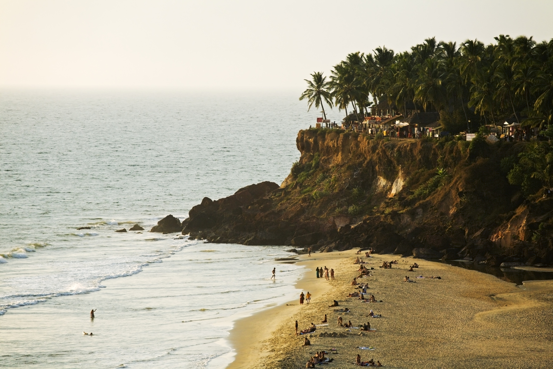 Tropical beach with people, palm trees, and cliffside cafe at sunset