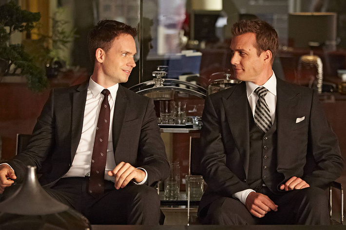 Two men in suits sit together looking cheerful, one with a drinks decanter nearby, in a scene suggesting a formal or professional setting