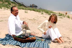 Two people laughing on a beach picnic, man in white shirt and trousers, woman in knit sweater and trousers