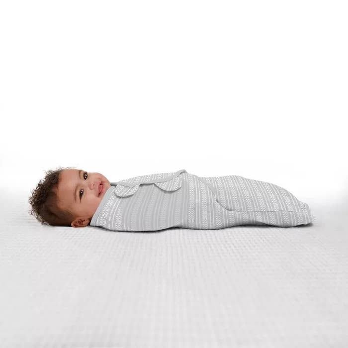 Baby rests in a grey wearable blanket on a plain background, looking to the side with a smile