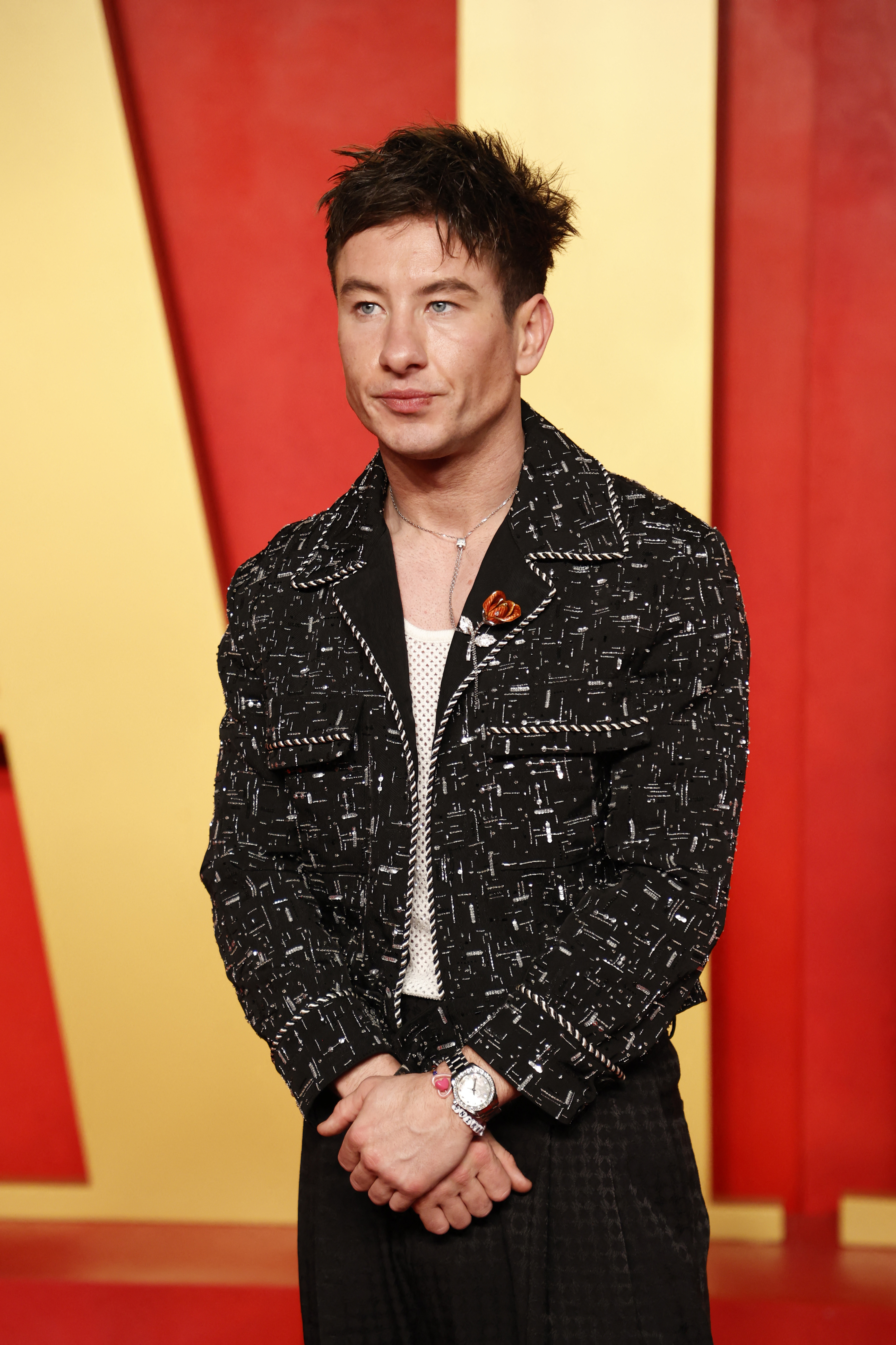 Barry Keoghan at an event wearing a patterned jacket with a red poppy pin and a watch, hands clasped in front