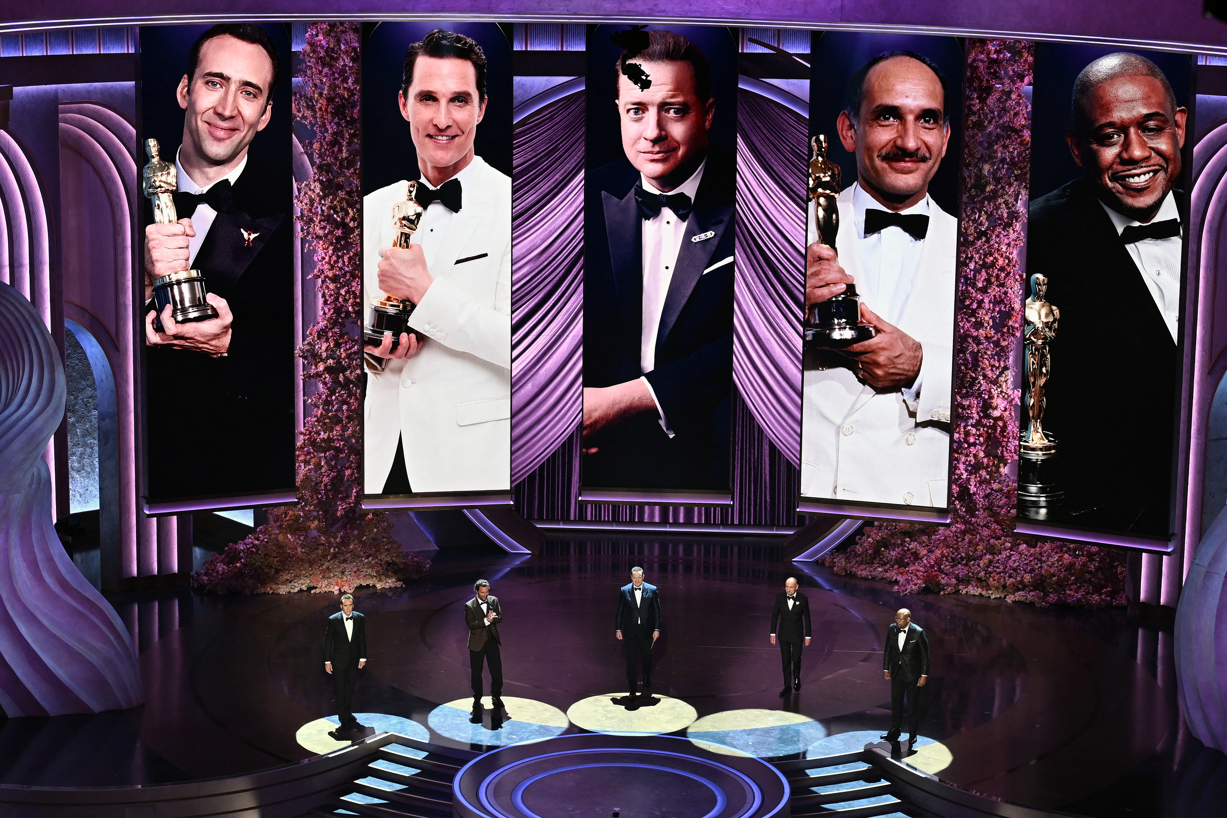 Five actors on stage at an awards show, each holding a trophy, with large portraits of them in the background