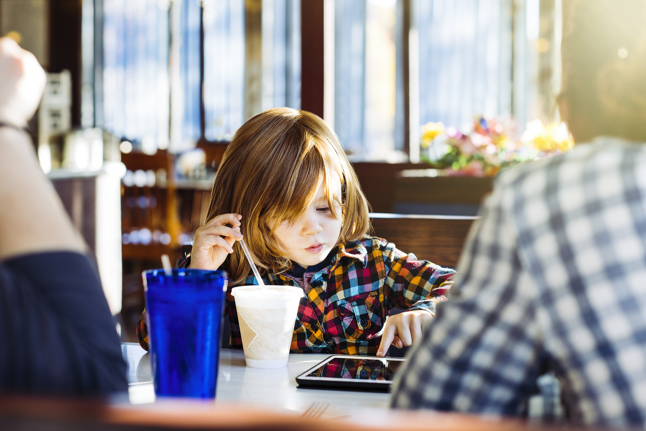 Child in plaid shirt focused on using a tablet at a restaurant table with an adult nearby