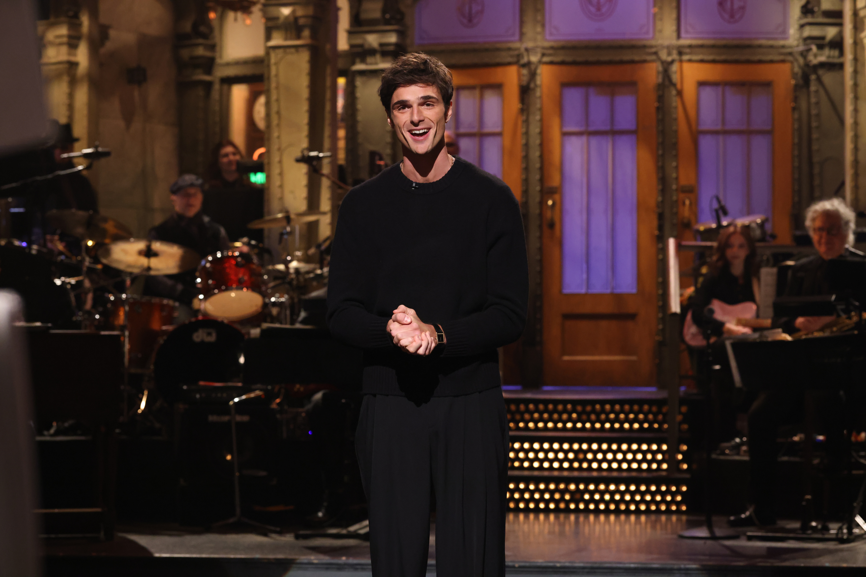 Jacob Elordi stands smiling on a stage with a band in the background during his SNL monologue