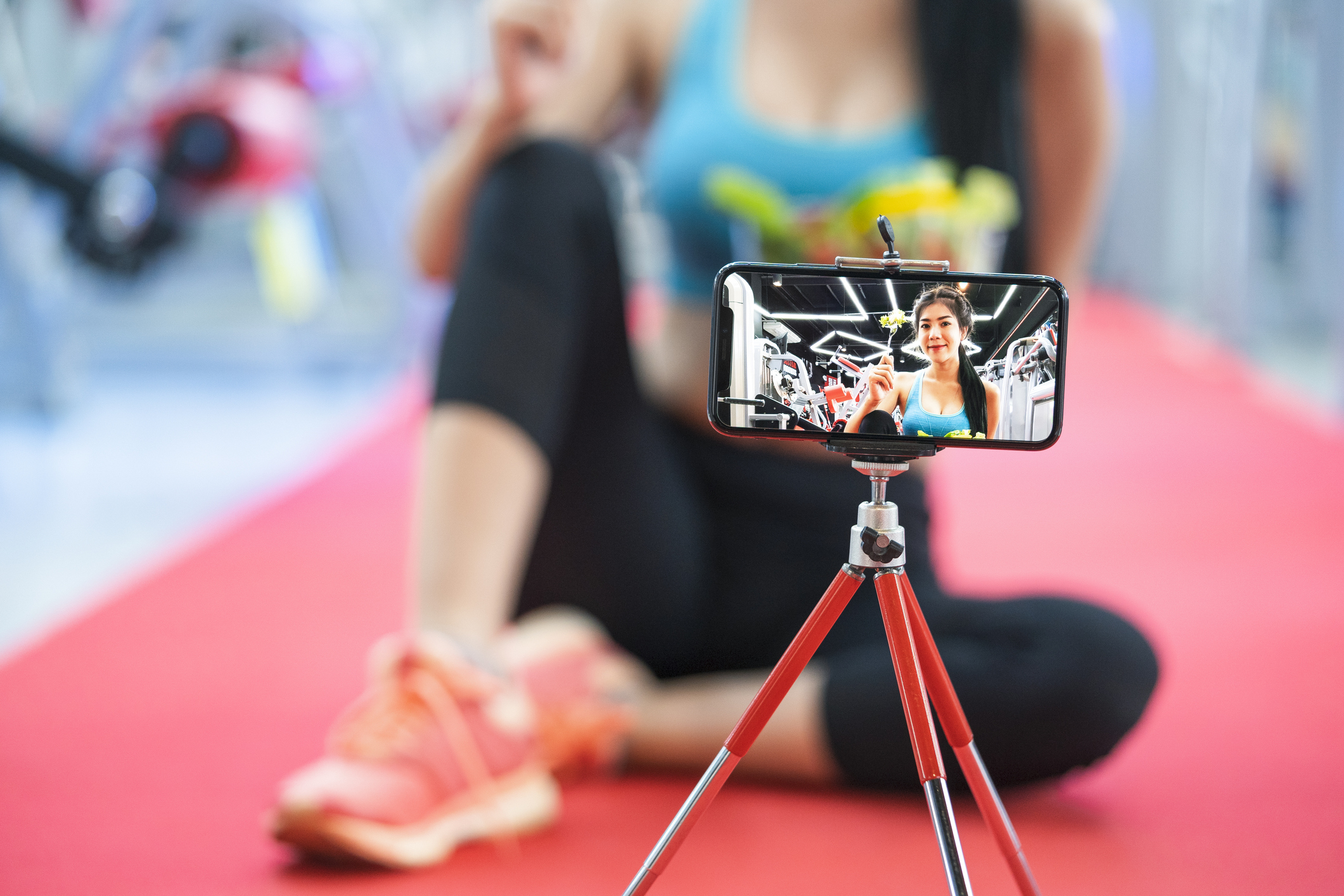 Woman records fitness vlog on smartphone with tripod, wearing workout attire, sitting on exercise mat