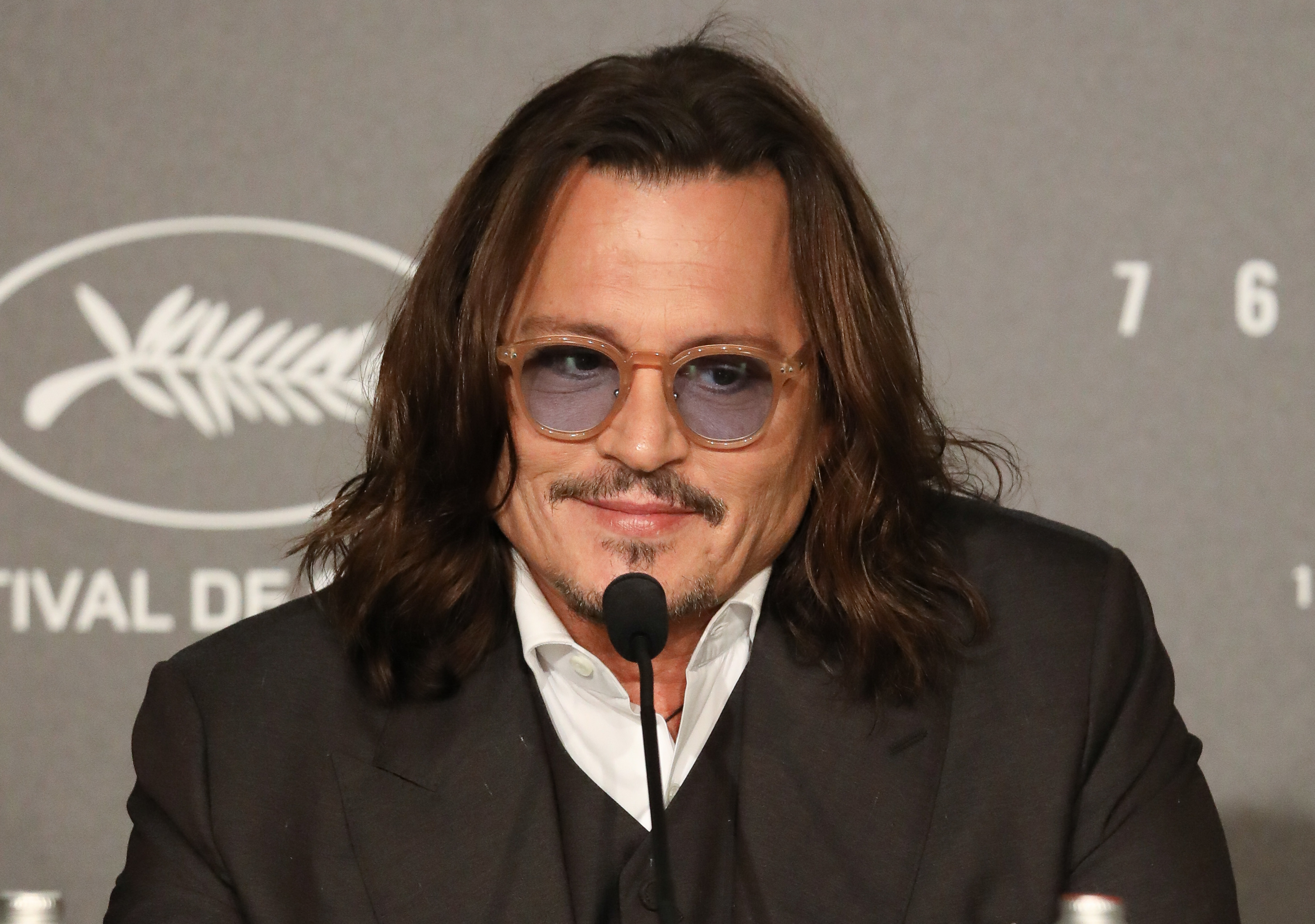 Johnny Depp wearing glasses and a suit, speaking into a microphone at an event