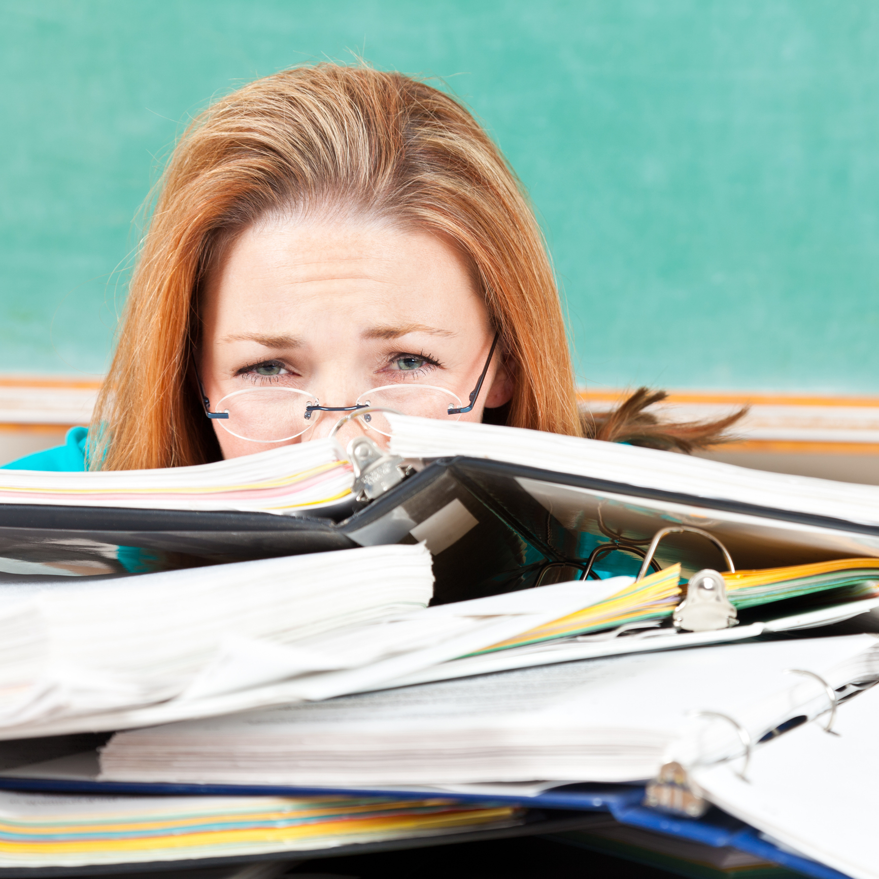 Woman with glasses peeking over a stack of paperwork on a desk, feeling overwhelmed