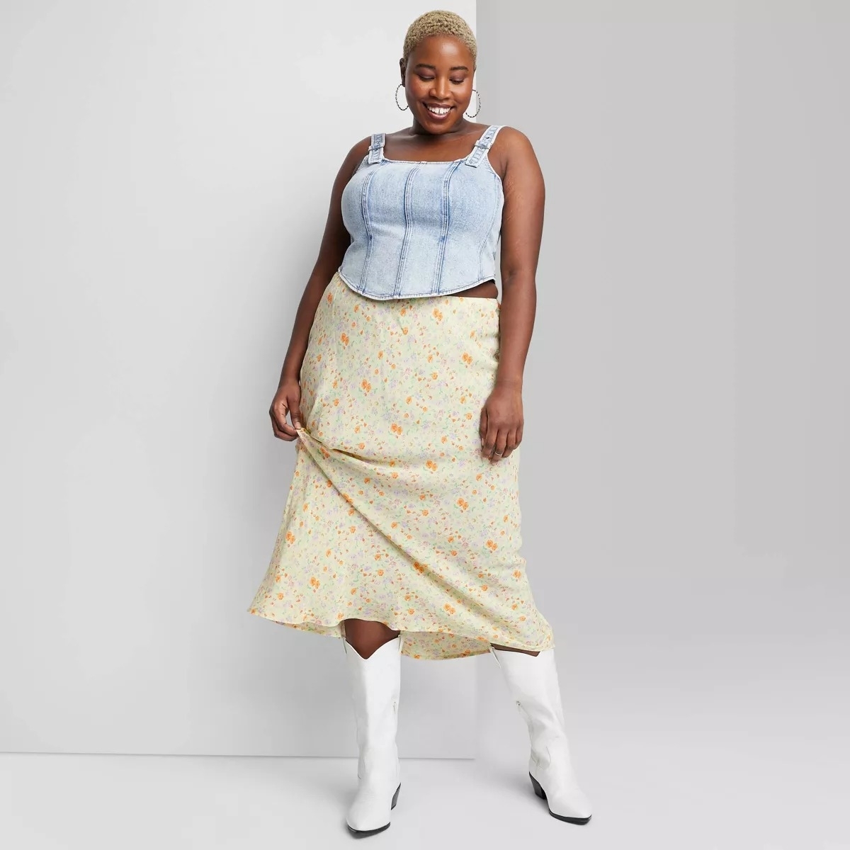 model in a sleeveless top, floral skirt, and white boots, posing with hand on skirt hem