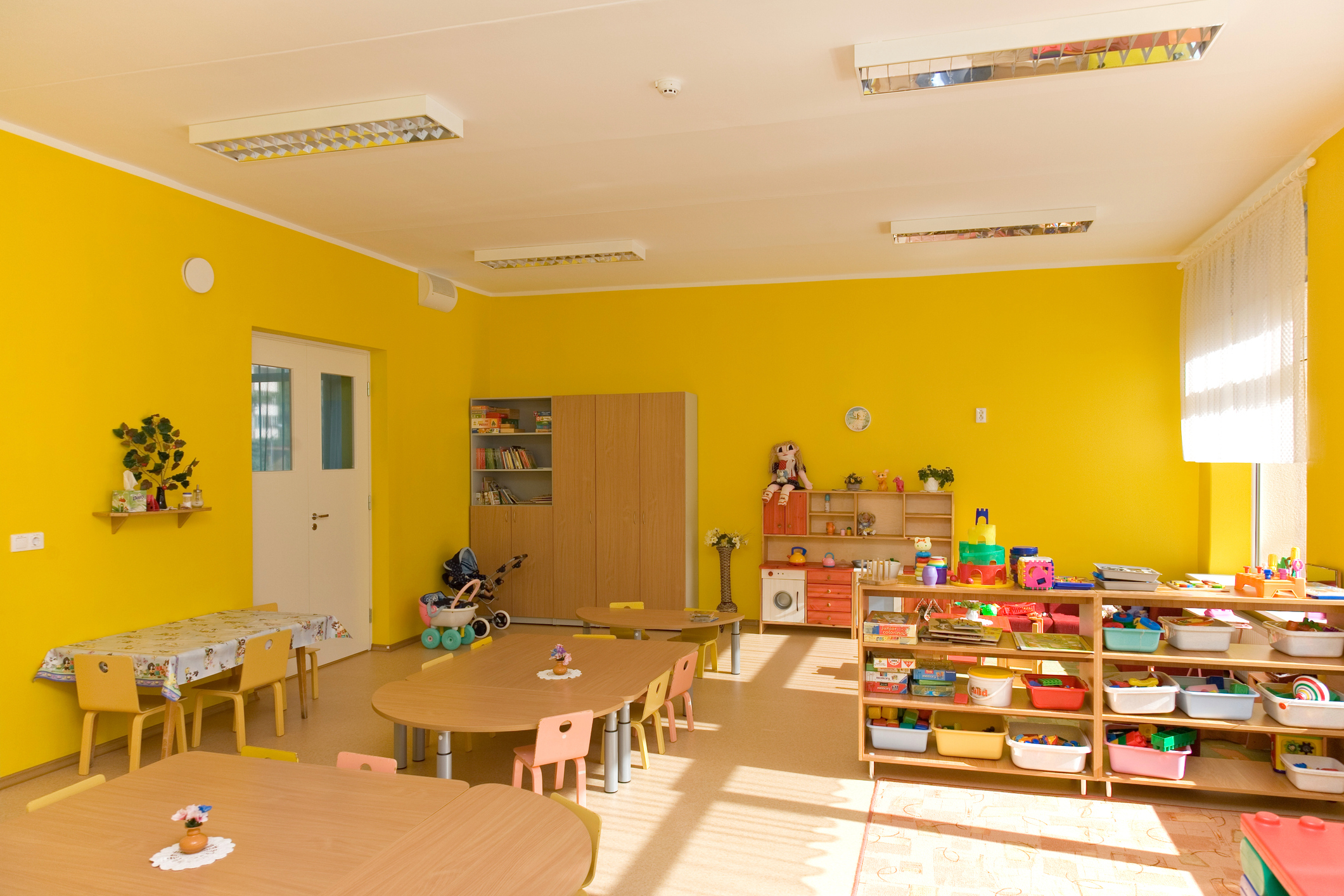 Brightly lit classroom with child-sized tables, chairs, and various educational toys and materials