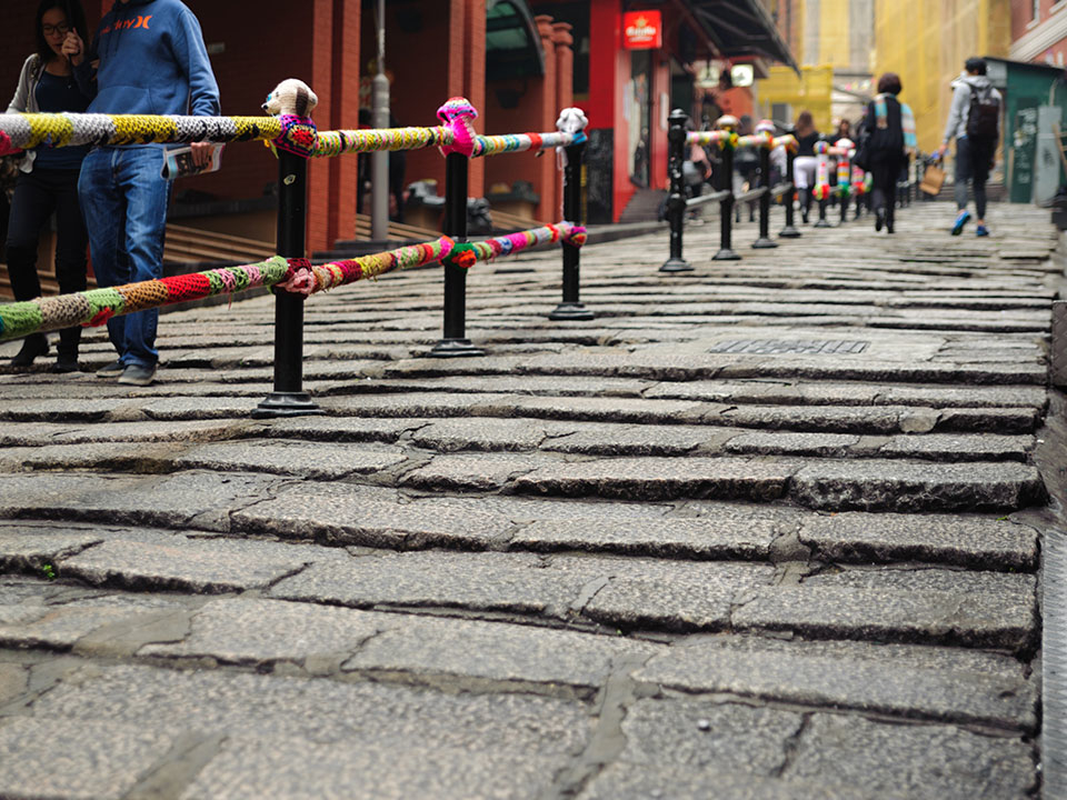 Pedestrians walk along a cobblestone street with yarn-bombed railings