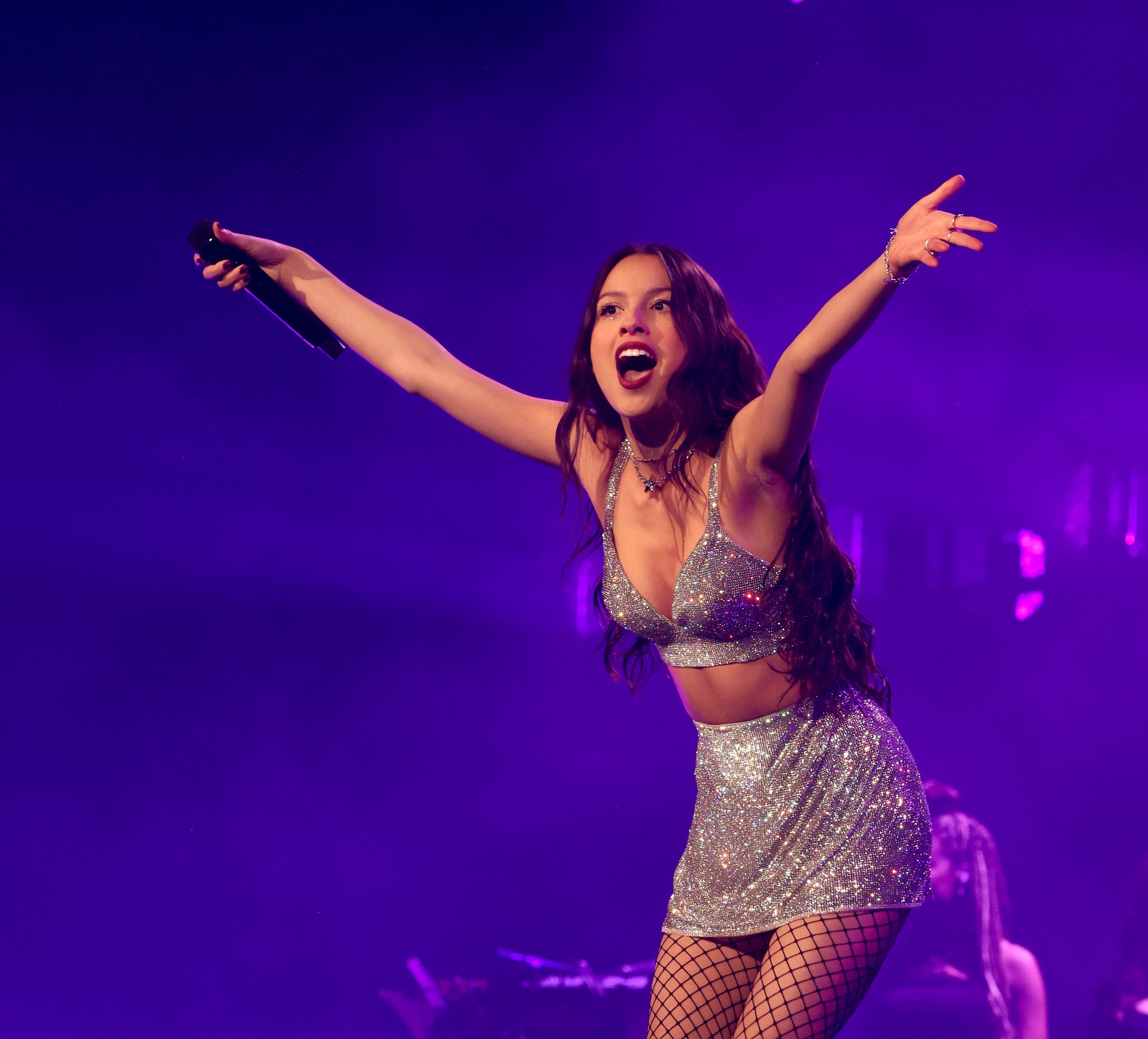 Olivia Rodrigo on stage in sparkly top and skirt, performing with her arms outstretched