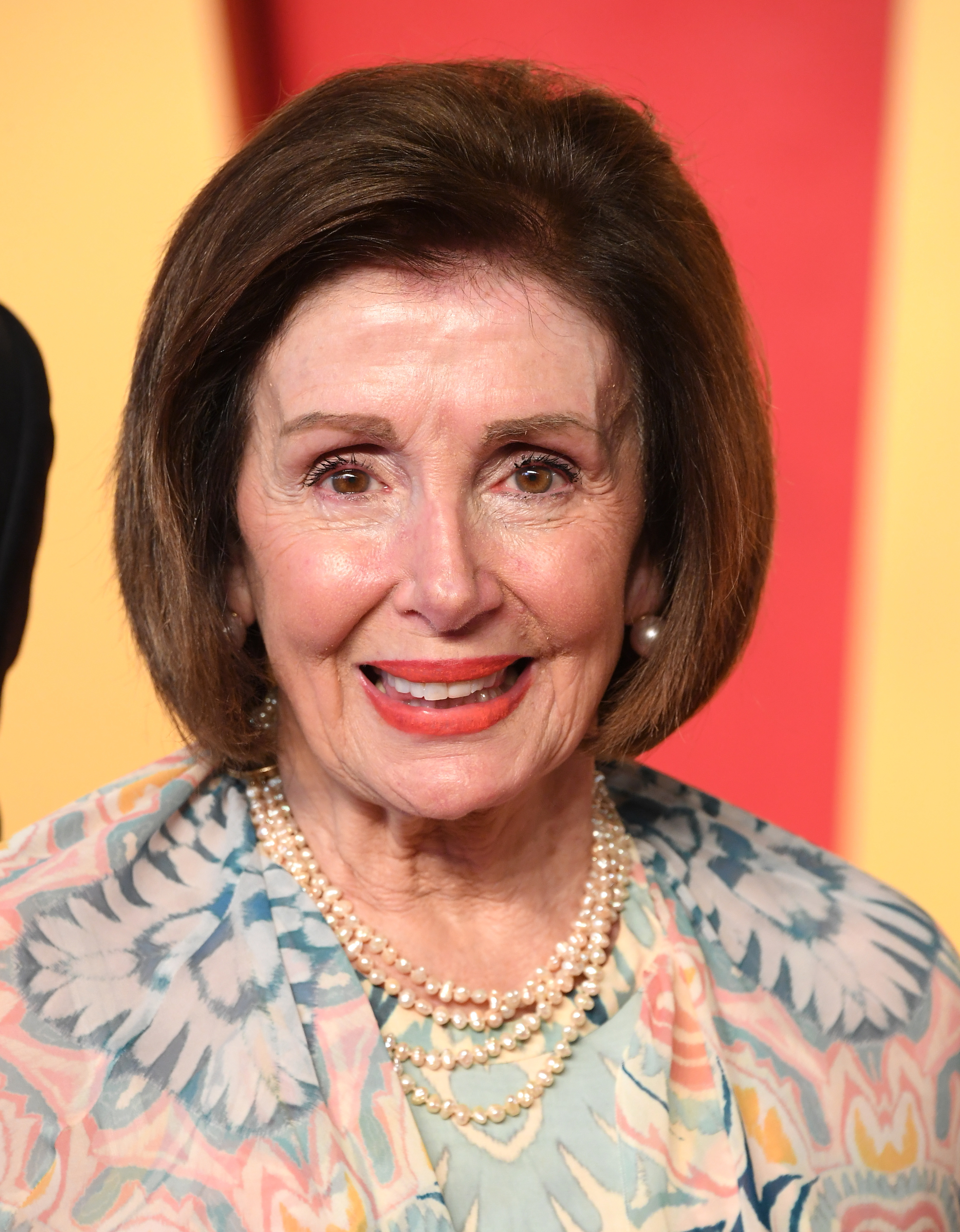 Nancy Pelosi smiling and wearing a patterned dress and pearl necklace