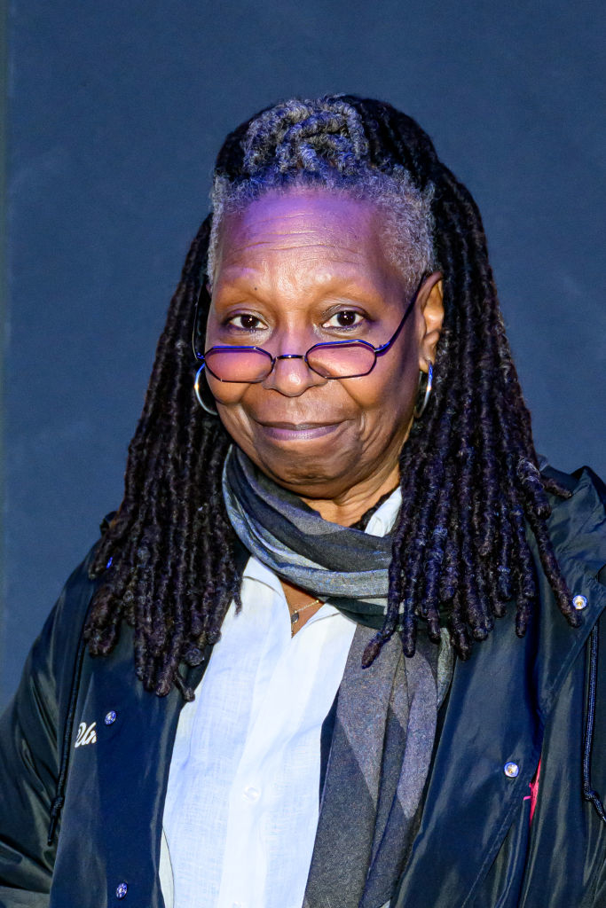 Whoopi Goldberg wearing glasses, a scarf, and a jacket with visible badges