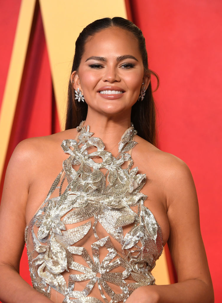 Chrissy Teigen in a detailed silver gown with a cut-out pattern, posing at an event