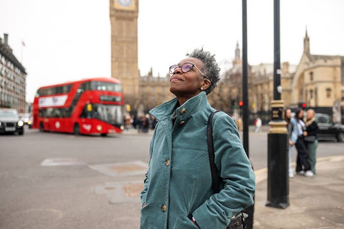 Person smiling with closed eyes, hands in coat pockets, Big Ben and red double-decker bus in background