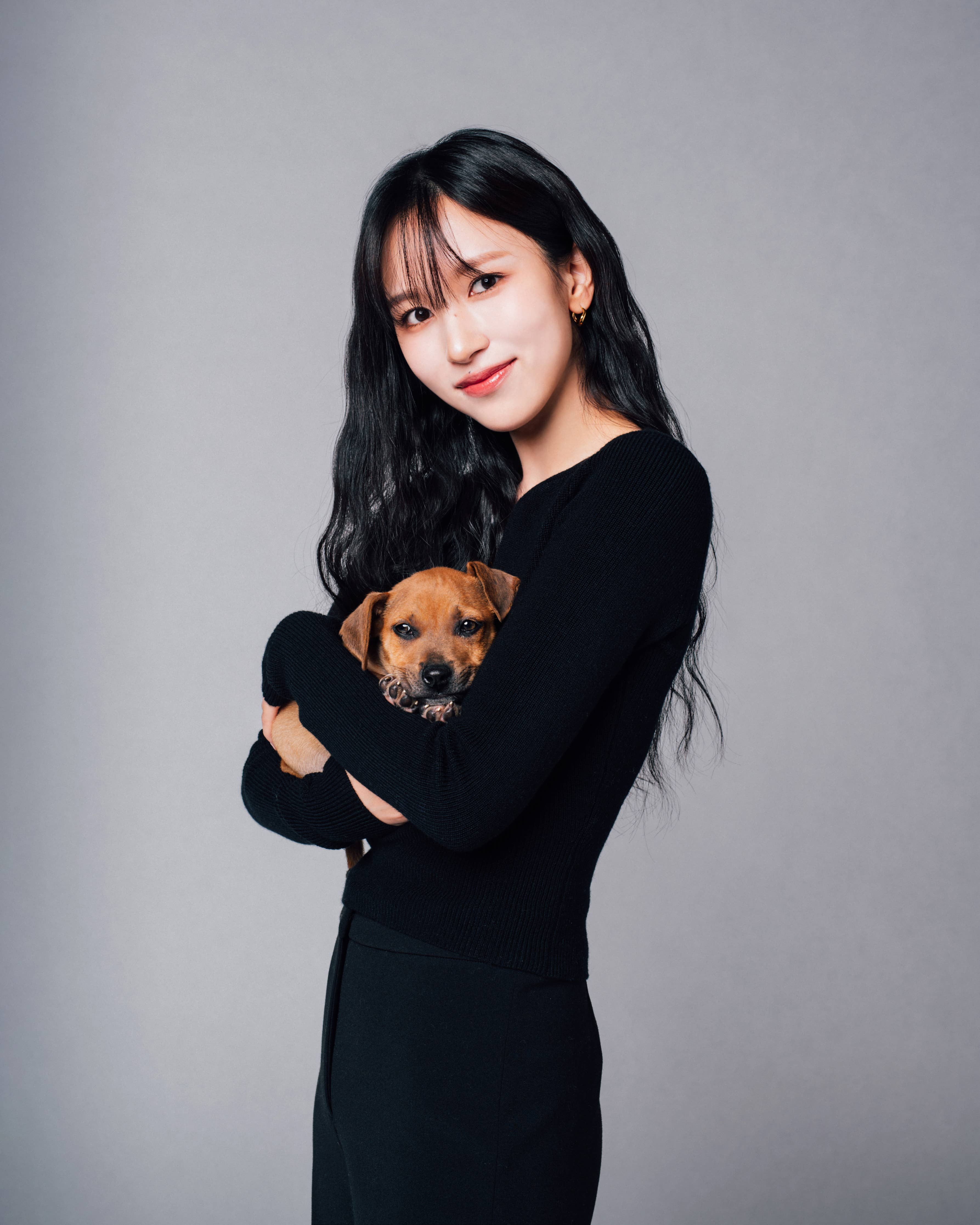 Woman in a black outfit smiling, holding a small puppy against a neutral background