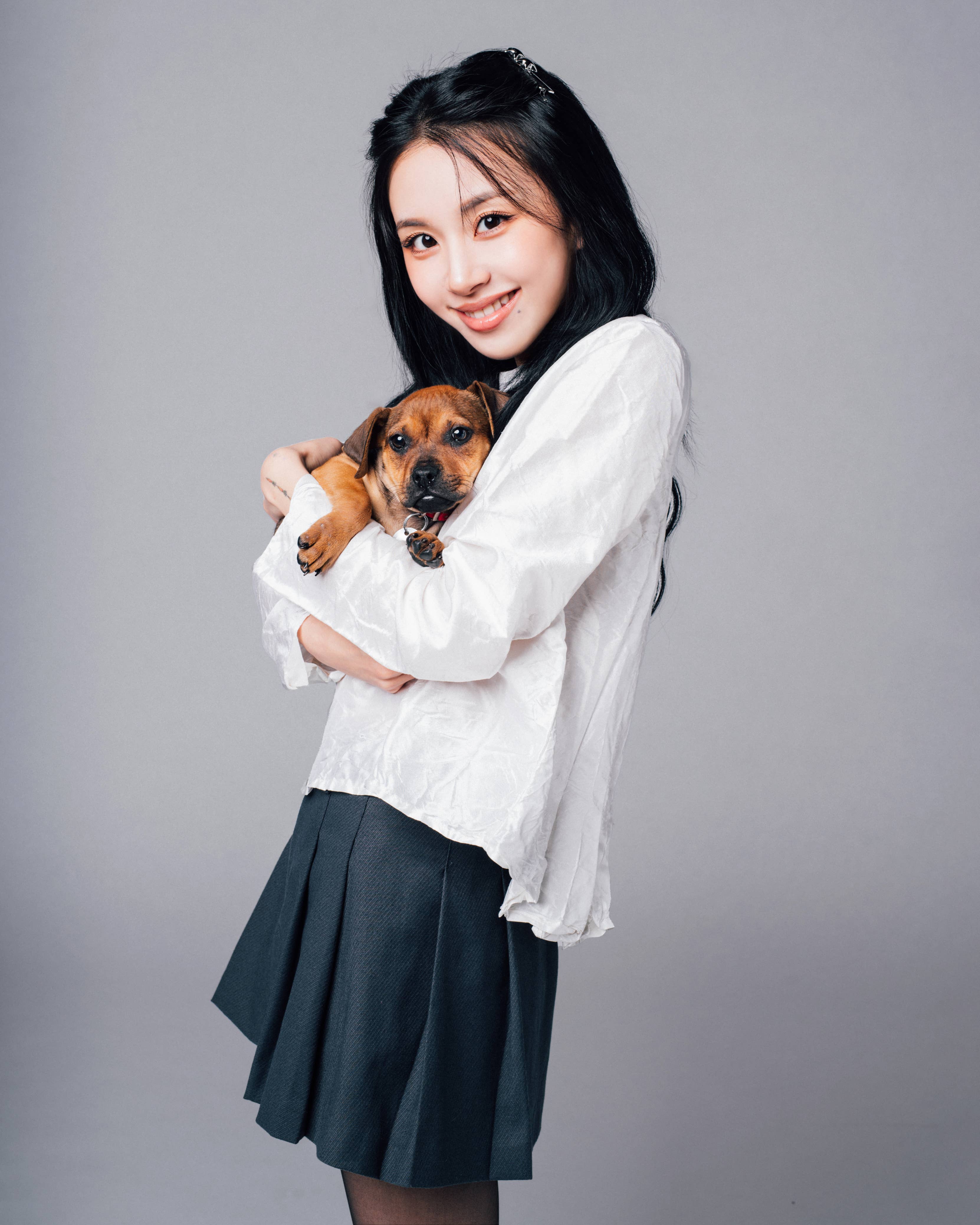 Woman holding a small dog, smiling at the camera, wearing a white blouse and black pleated skirt