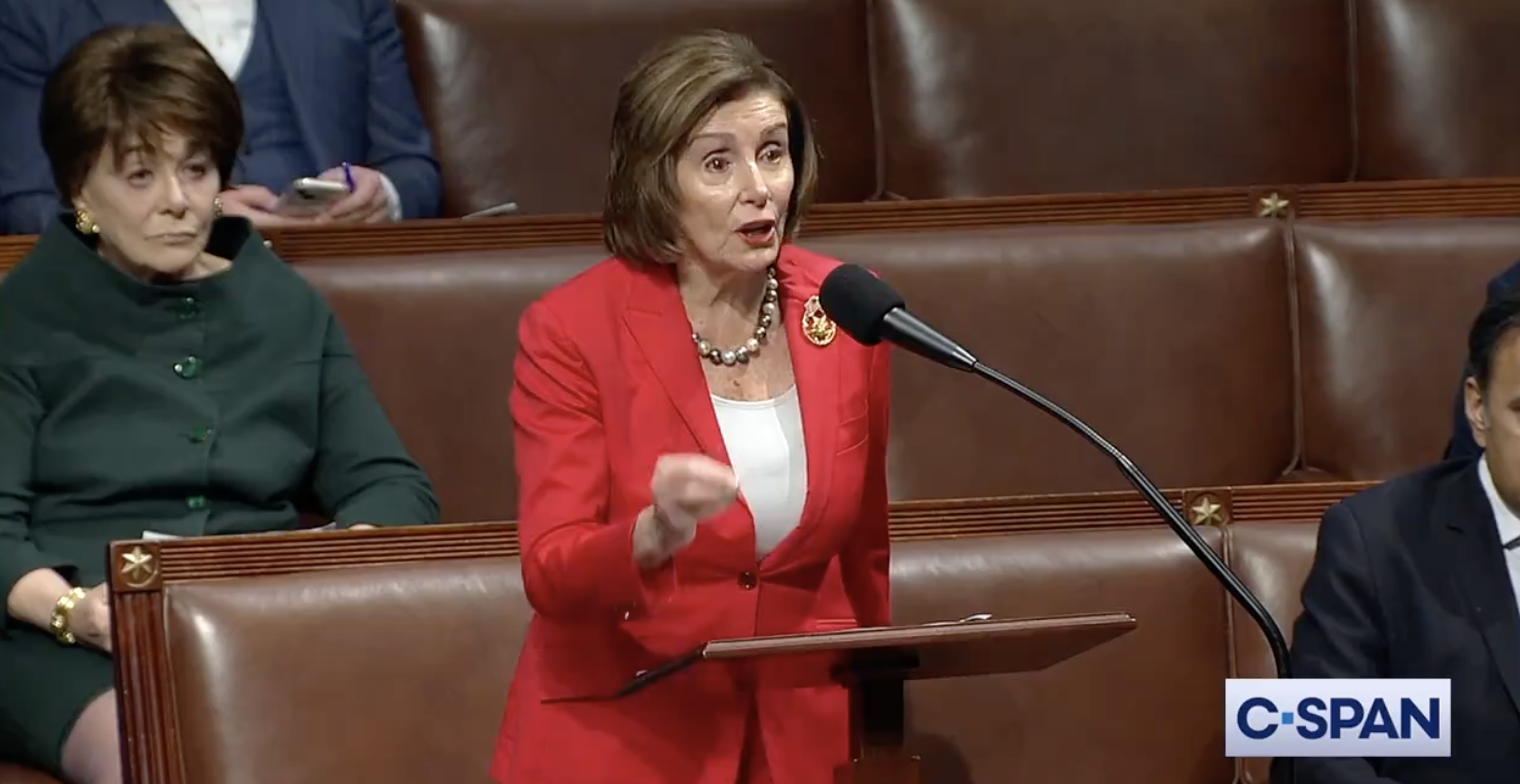 Nancy Pelosi in red speaking at a podium in the House of Representatives chamber
