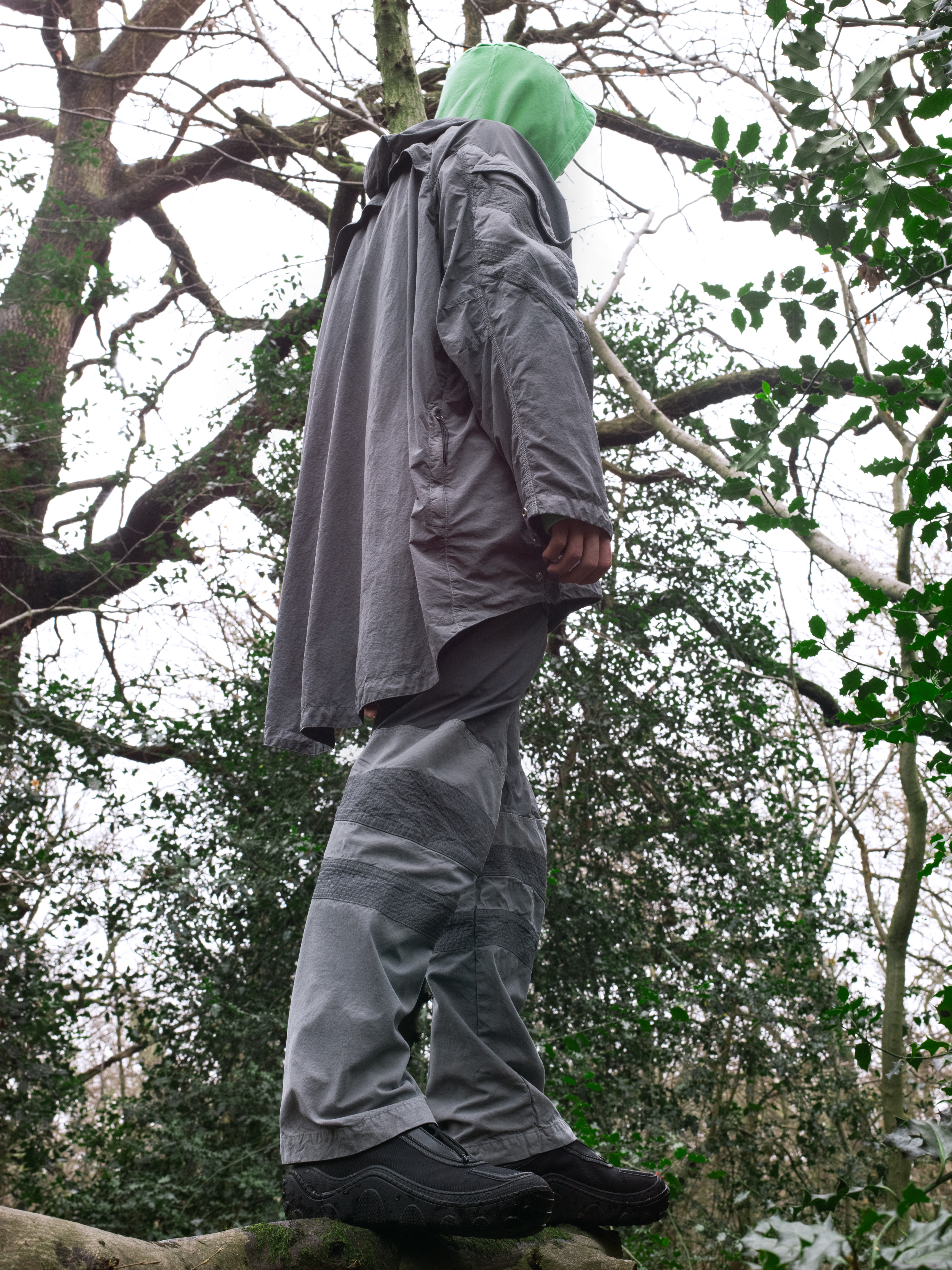 Person in outdoor clothing stands on a tree trunk, face obscured by a hood, amidst greenery