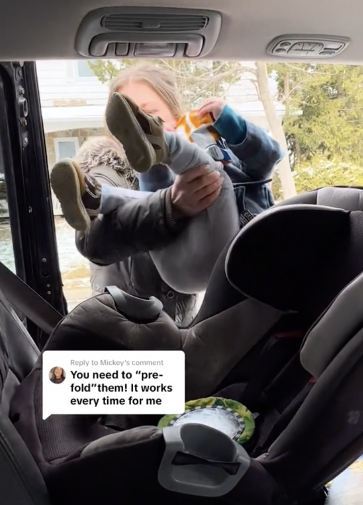 Person struggles to put child&#x27;s legs into a car seat as the child leans backward outside the car