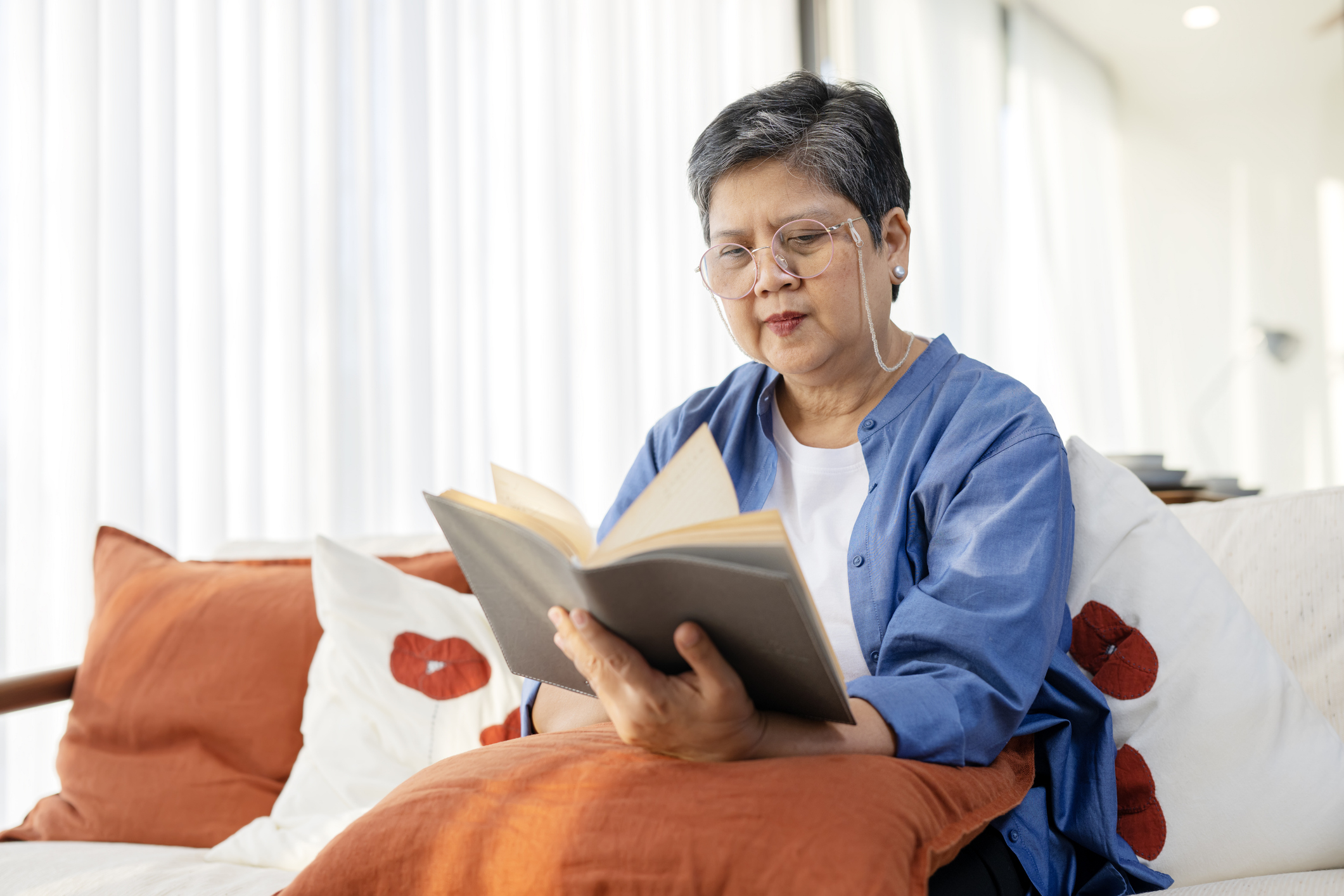Woman in glasses reading a book on a couch with decorative pillows